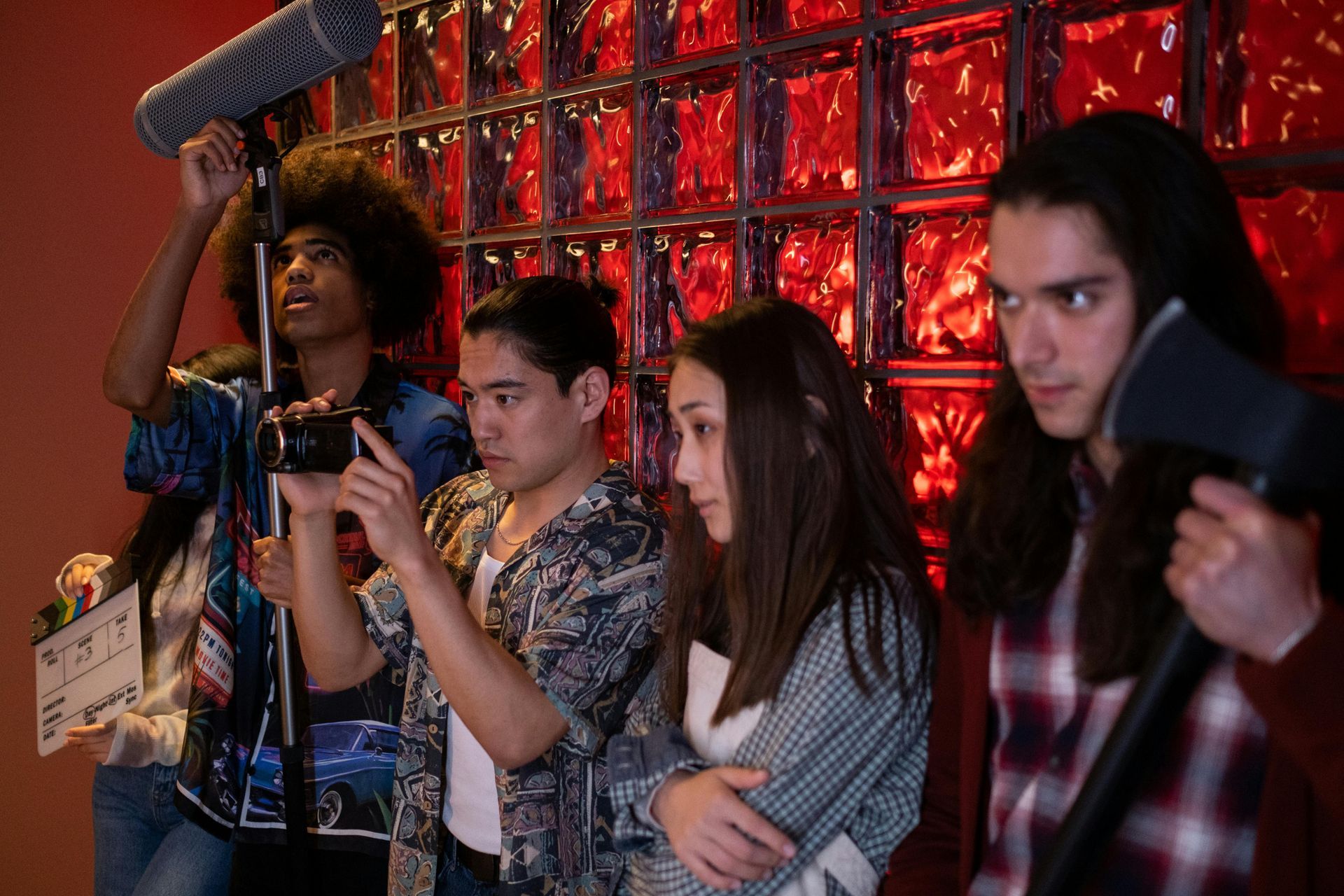 Film crew on set; young people with camera, boom mic, clapperboard, axe, and worried expressions. Brick backdrop.