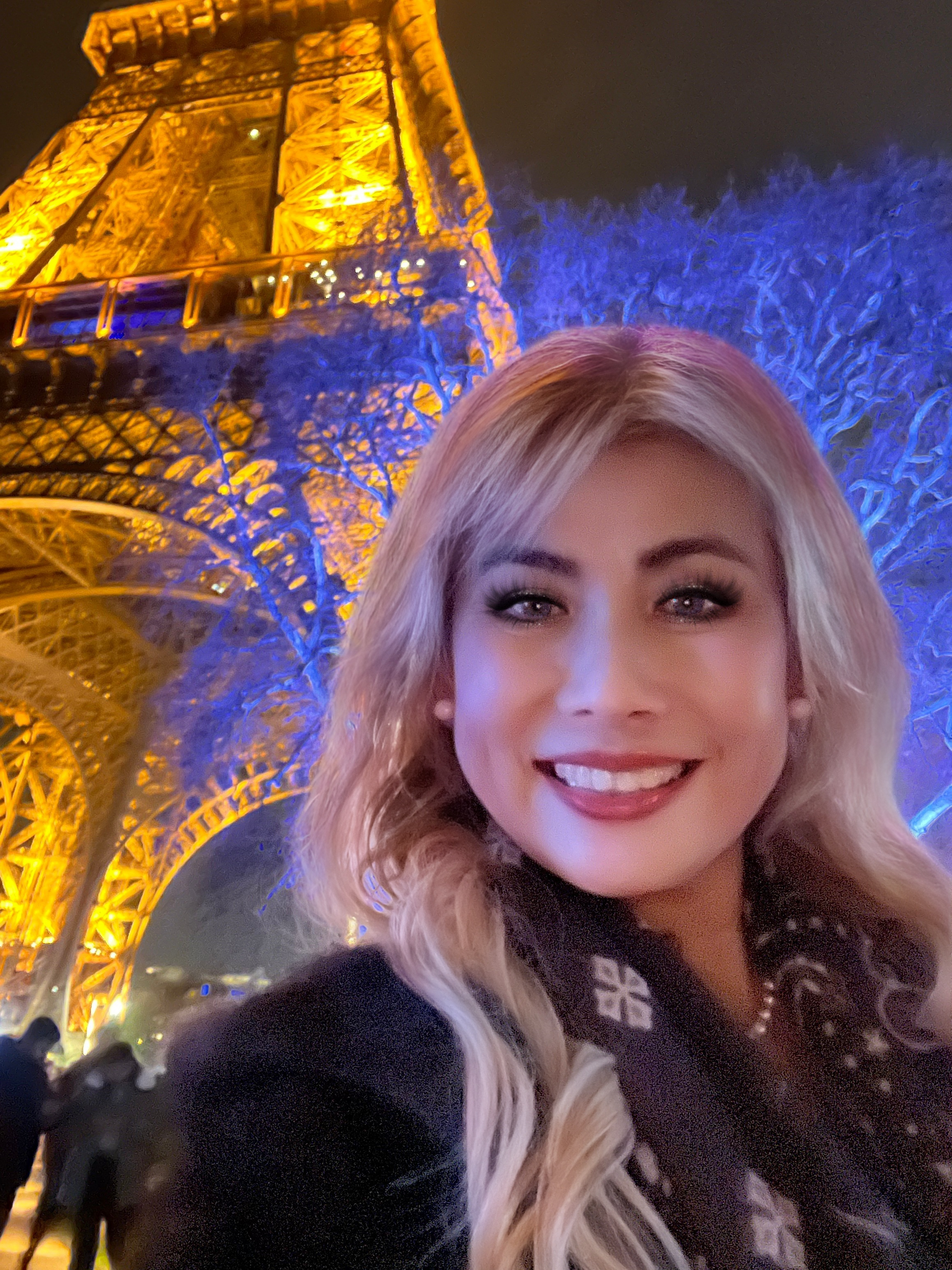 Woman smiling in front of the Eiffel Tower, illuminated with blue and gold lights at night.