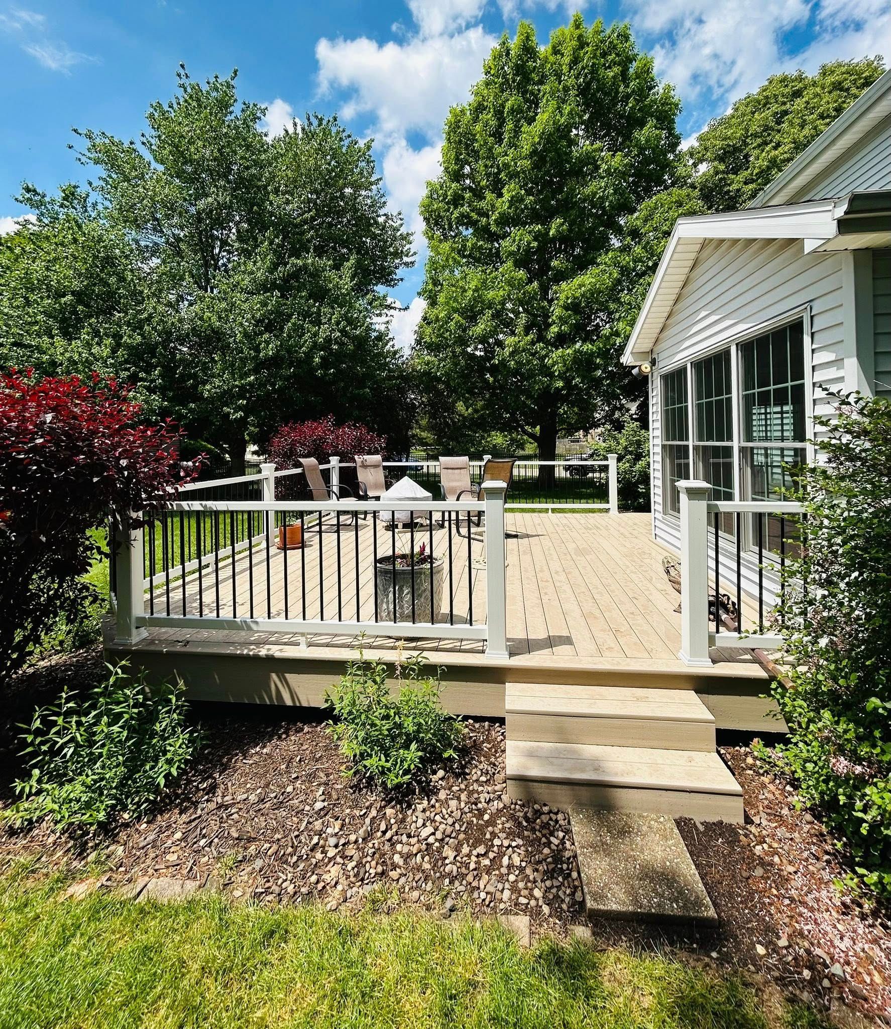 Wooden deck with white railing and steps, attached to a house with a screened porch. Green trees and bushes surround the deck.