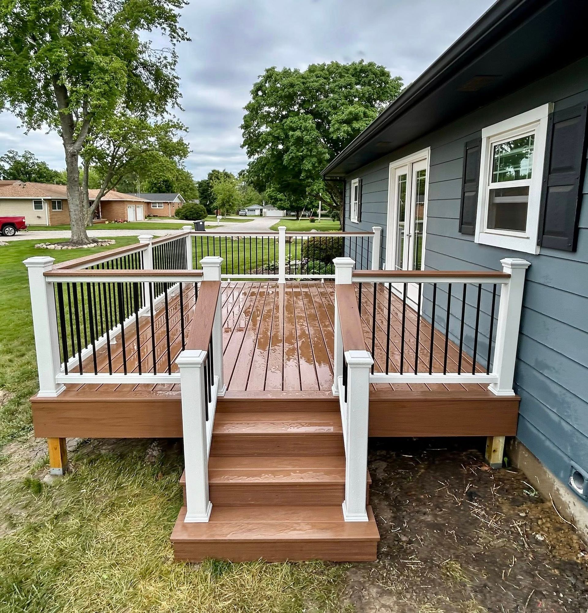A newly built wooden deck with brown planks, white posts, and black railings, attached to a blue house. Stairs lead down to the yard.