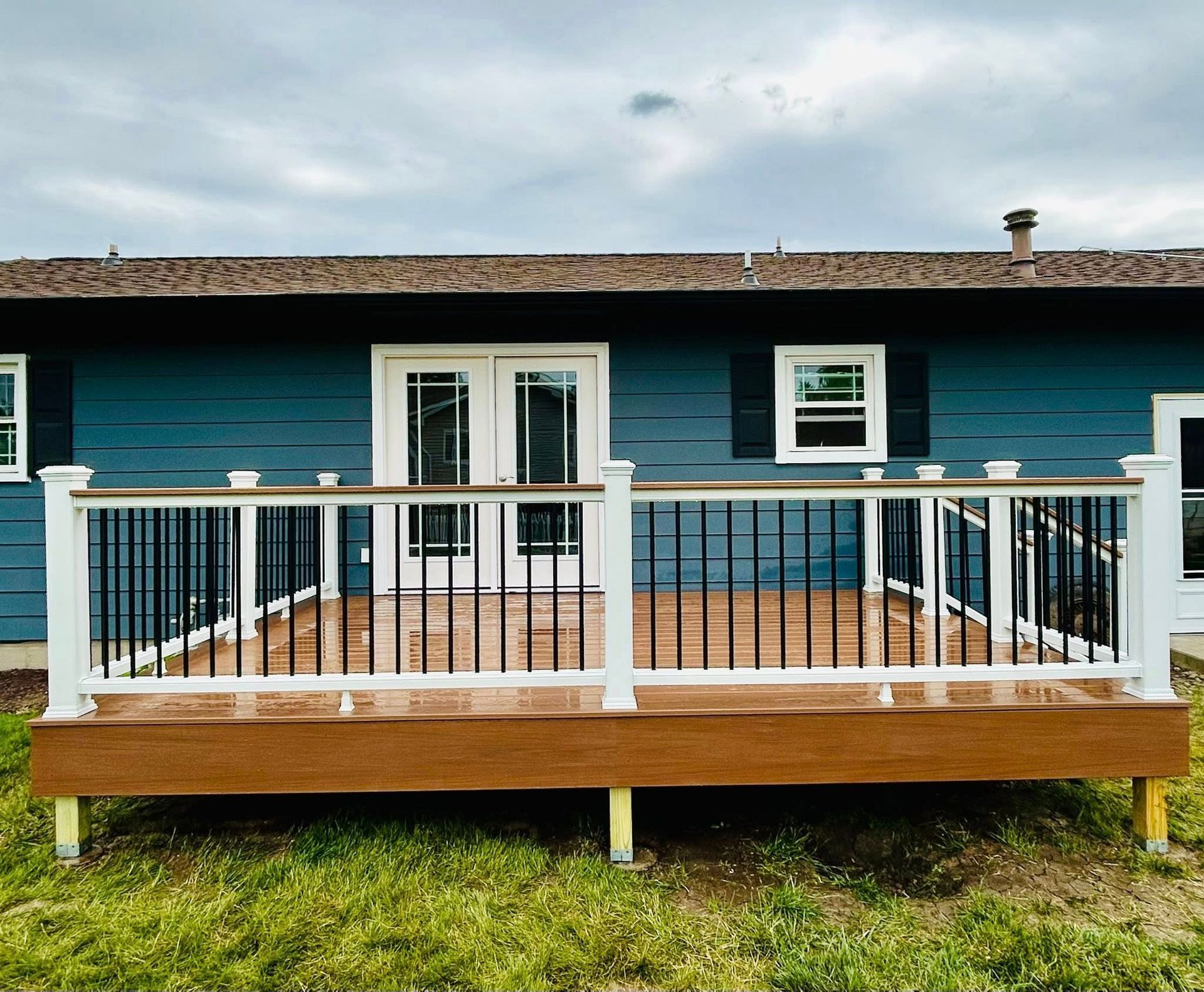 A wooden deck with white railings and black spindles extends from a blue house with French doors.
