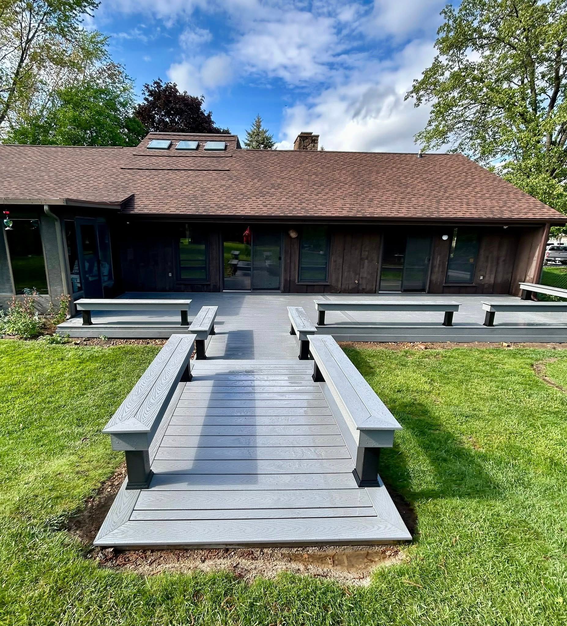 Gray wooden walkway with benches leading to a brown-roofed house with glass doors and windows, set on green grass under a blue sky.