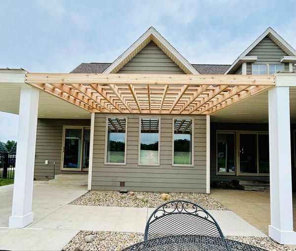 A wooden pergola above a concrete patio extends from a house with a gray exterior, three windows, and white columns.