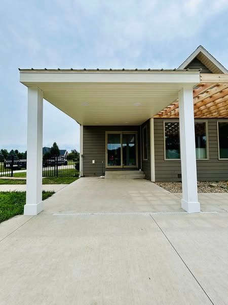 Exterior view of a house with a concrete patio and a covered seating area supported by white columns. The siding is grey and a sliding glass door is visible.