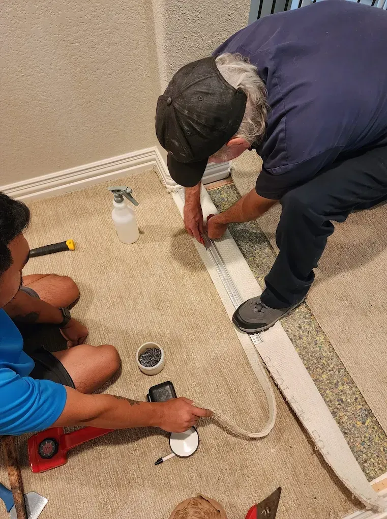 Two people installing carpet: one using a tool and the other guiding the carpet along the wall.
