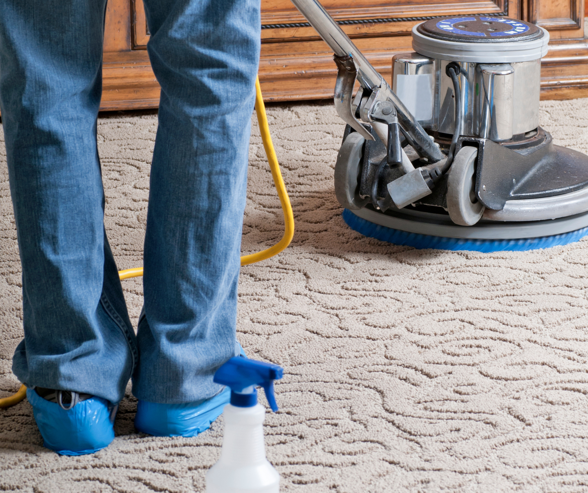 Person in jeans and blue shoe covers using a floor cleaning machine on a patterned carpet; spray bottle nearby.