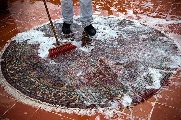 Person using a broom to scrub a wet, patterned rug on a tiled surface, surrounded by suds.