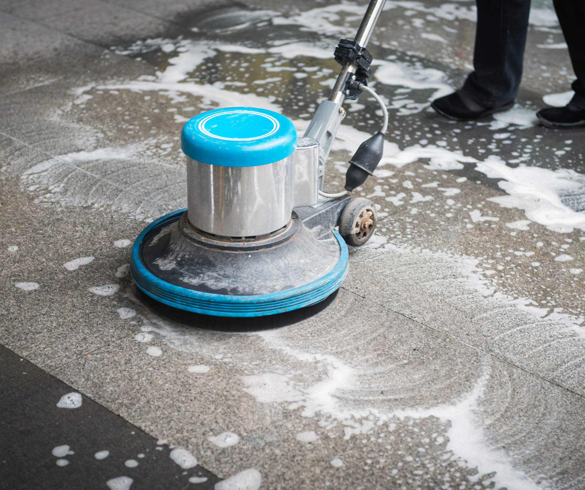 Floor cleaning machine scrubbing a stone surface, creating suds. Person's legs and shoes visible.