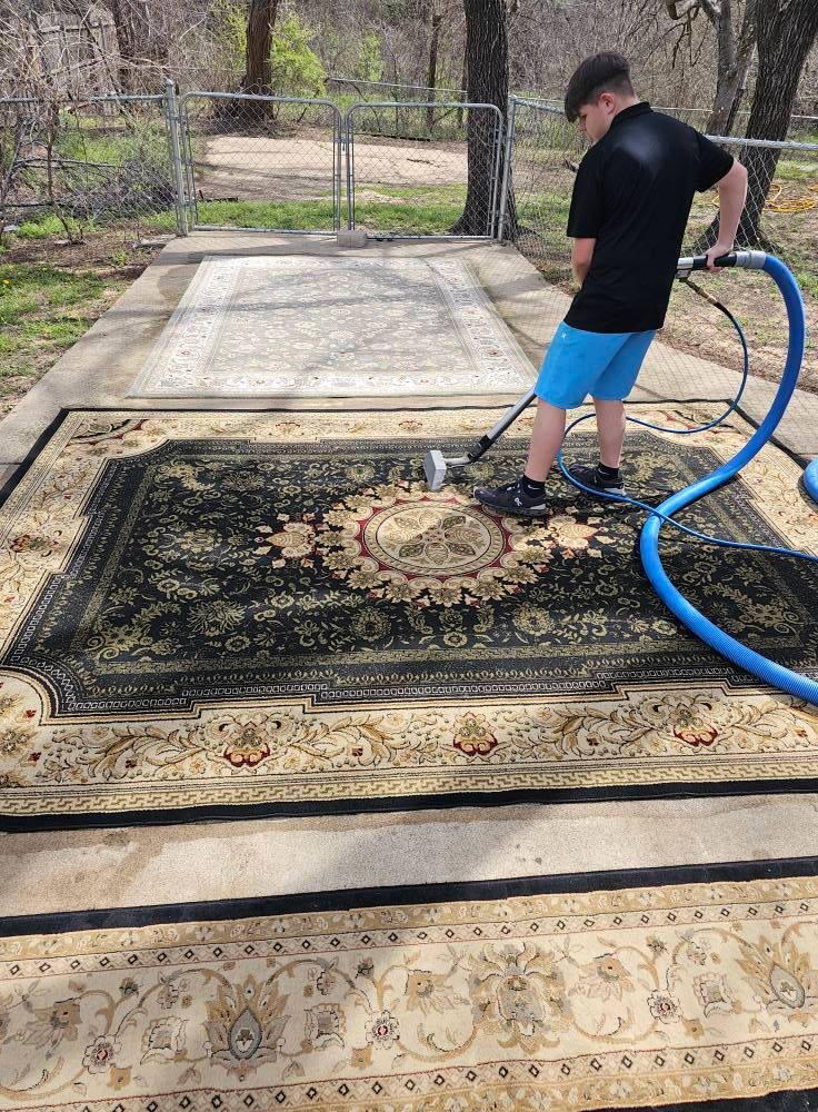 Person using a hose to clean a large black and beige rug outdoors on a concrete pad.