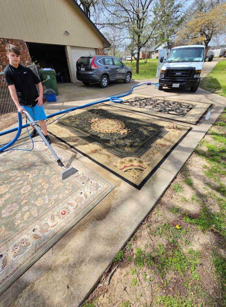 A person cleaning rugs outdoors with a machine. Three rugs laid out on concrete; van and vehicles in background.
