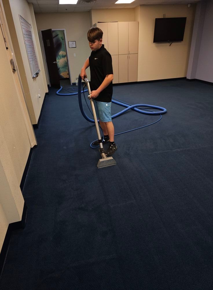 Person cleans a dark blue carpet with an extraction machine in a room with white walls and cabinets.