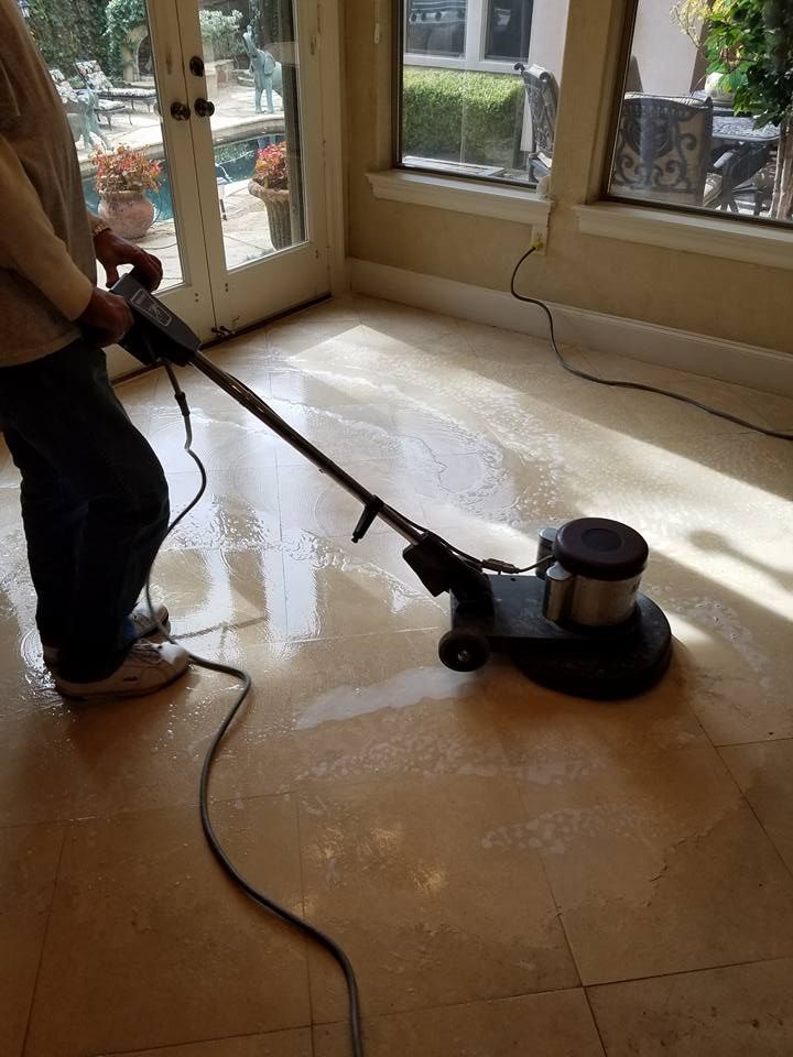 Person using a floor polishing machine on a light-colored tiled floor near a glass door and window.