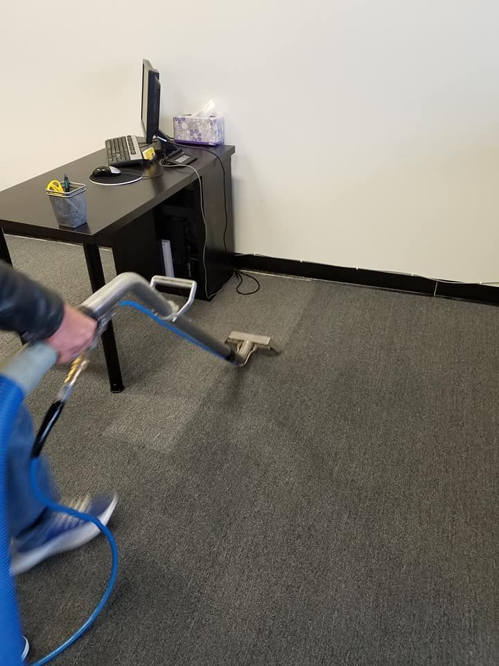 Person cleaning a grey office carpet with a blue cleaning machine. A black desk is nearby.