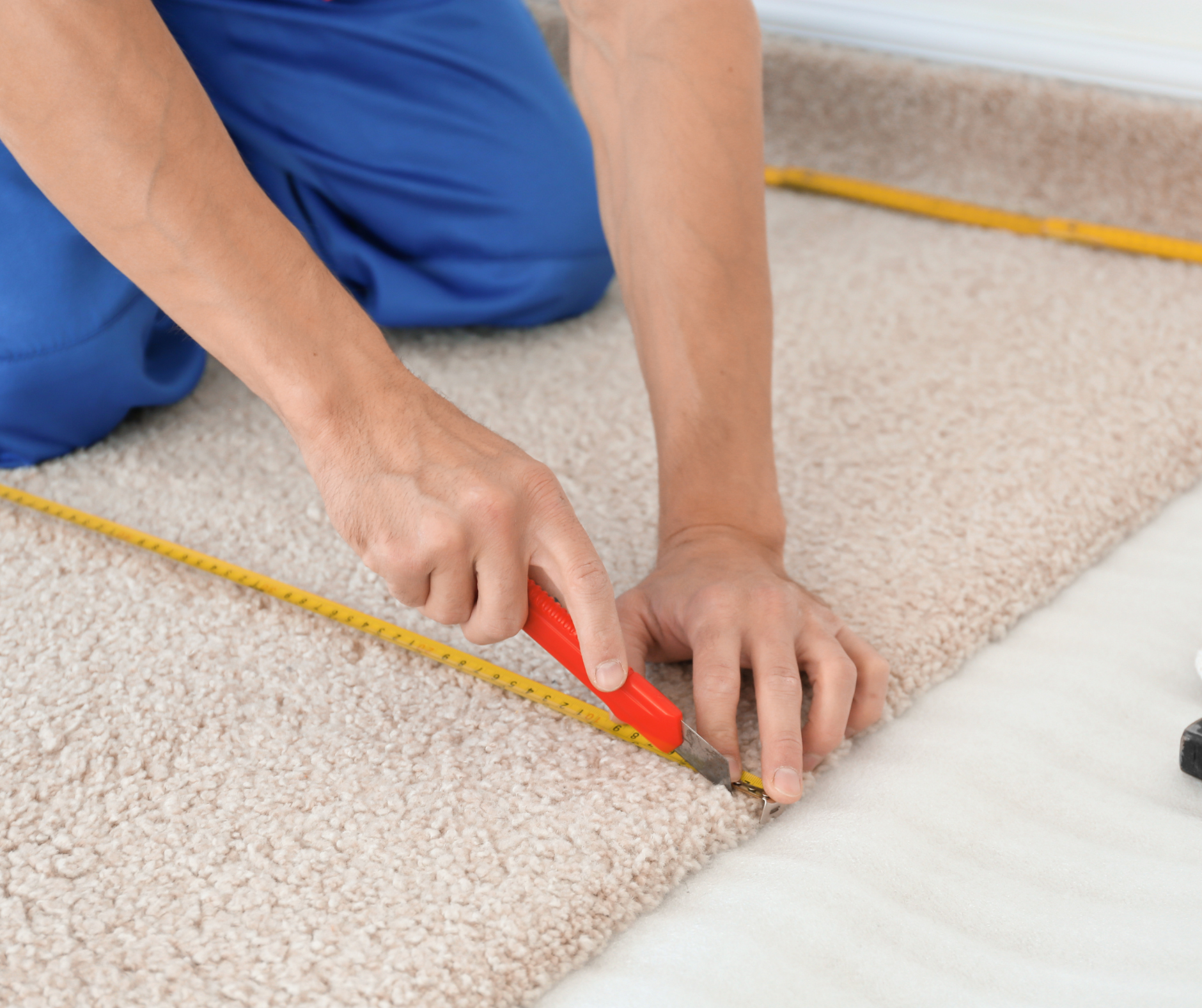 Person kneeling, cutting beige carpet with a utility knife and measuring tape.