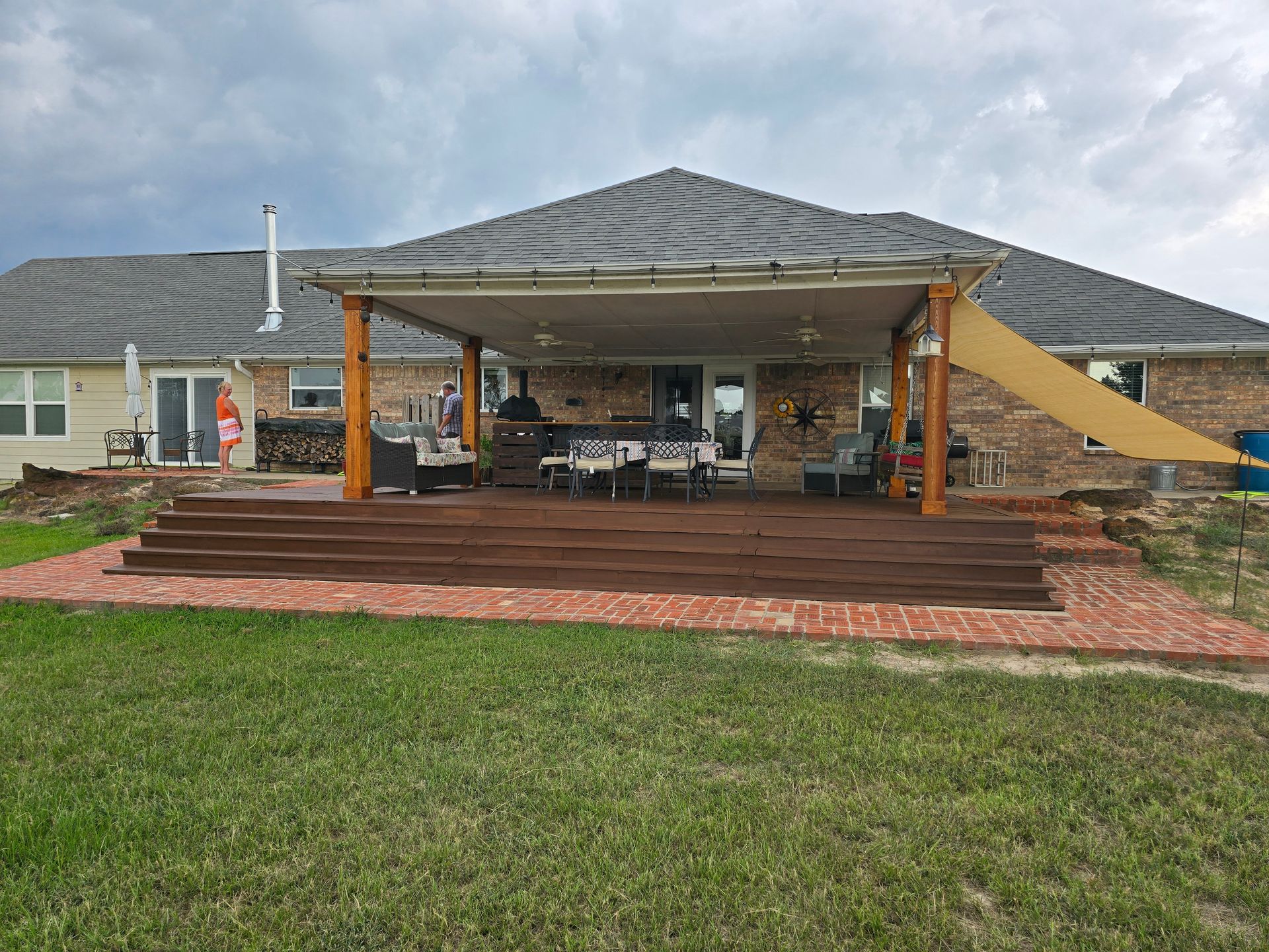 Backyard deck with awning, brick patio, and grass. Wooden deck steps lead to covered dining area; cloudy sky.