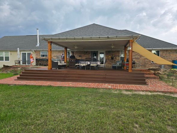 Backyard deck with awning, brick patio, and grass. Wooden deck steps lead to covered dining area; cloudy sky.