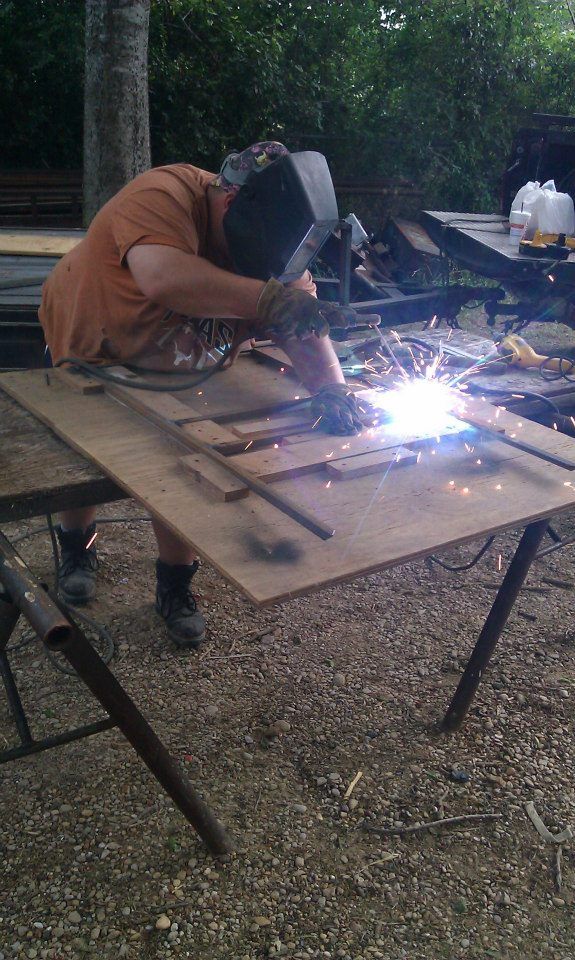 Welder in orange shirt and protective mask working on metal frame outdoors, sparks flying.