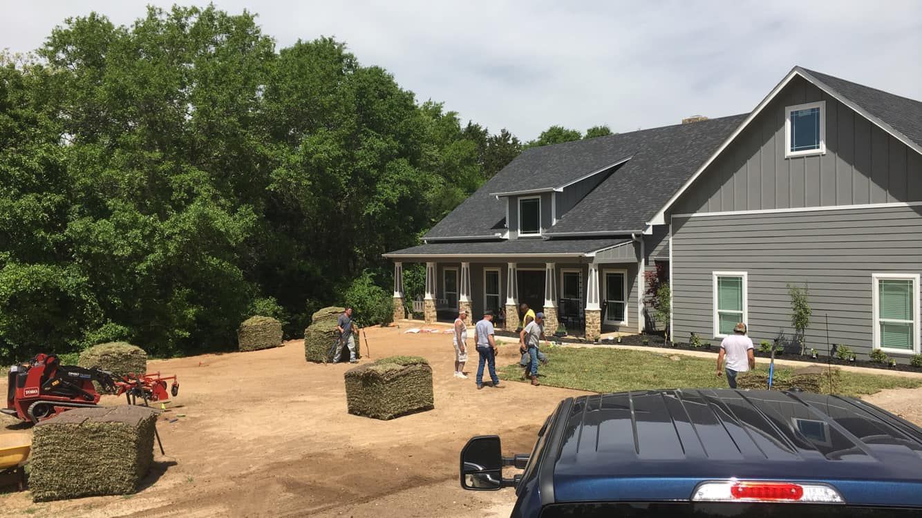 People laying sod near a gray house with a dark roof; hay bales and a tractor are nearby.