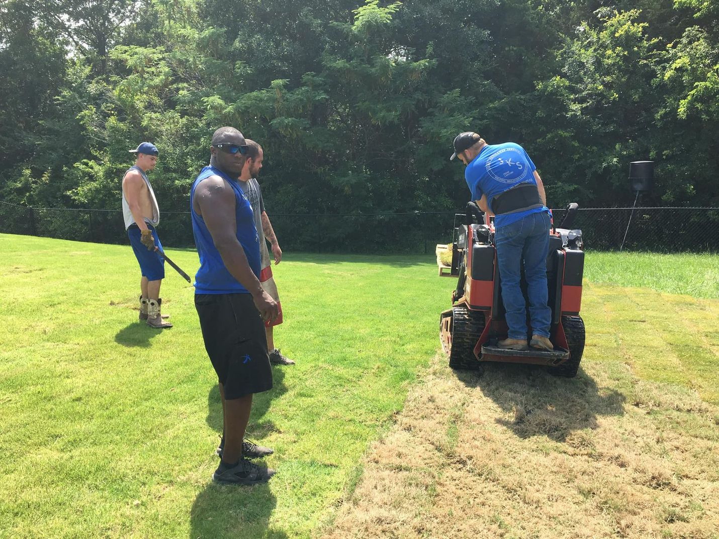 Four men on a lawn; one operates a small trencher, the others watch. Green grass, sunny day.