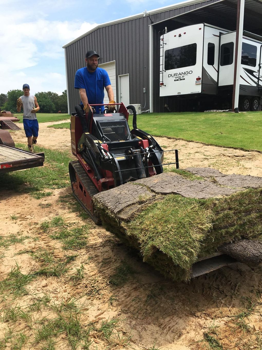 Man operating a mini-skid steer transporting sod. Another man walks nearby. Shed, RV, and outdoors.