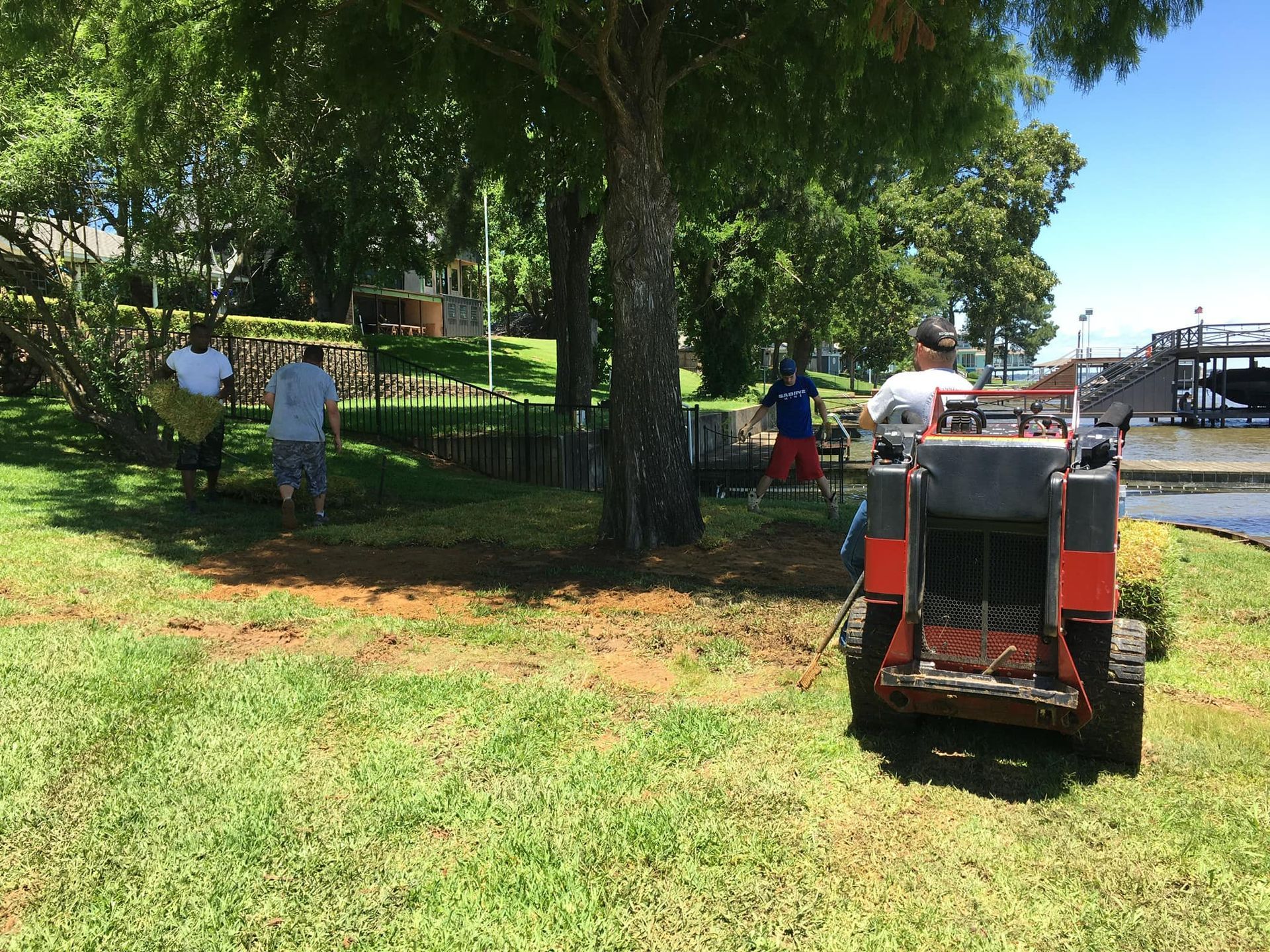 People working on lawn care by a waterfront, operating machinery, on a sunny day.