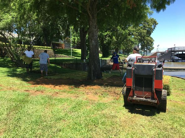 People working on lawn care by a waterfront, operating machinery, on a sunny day.