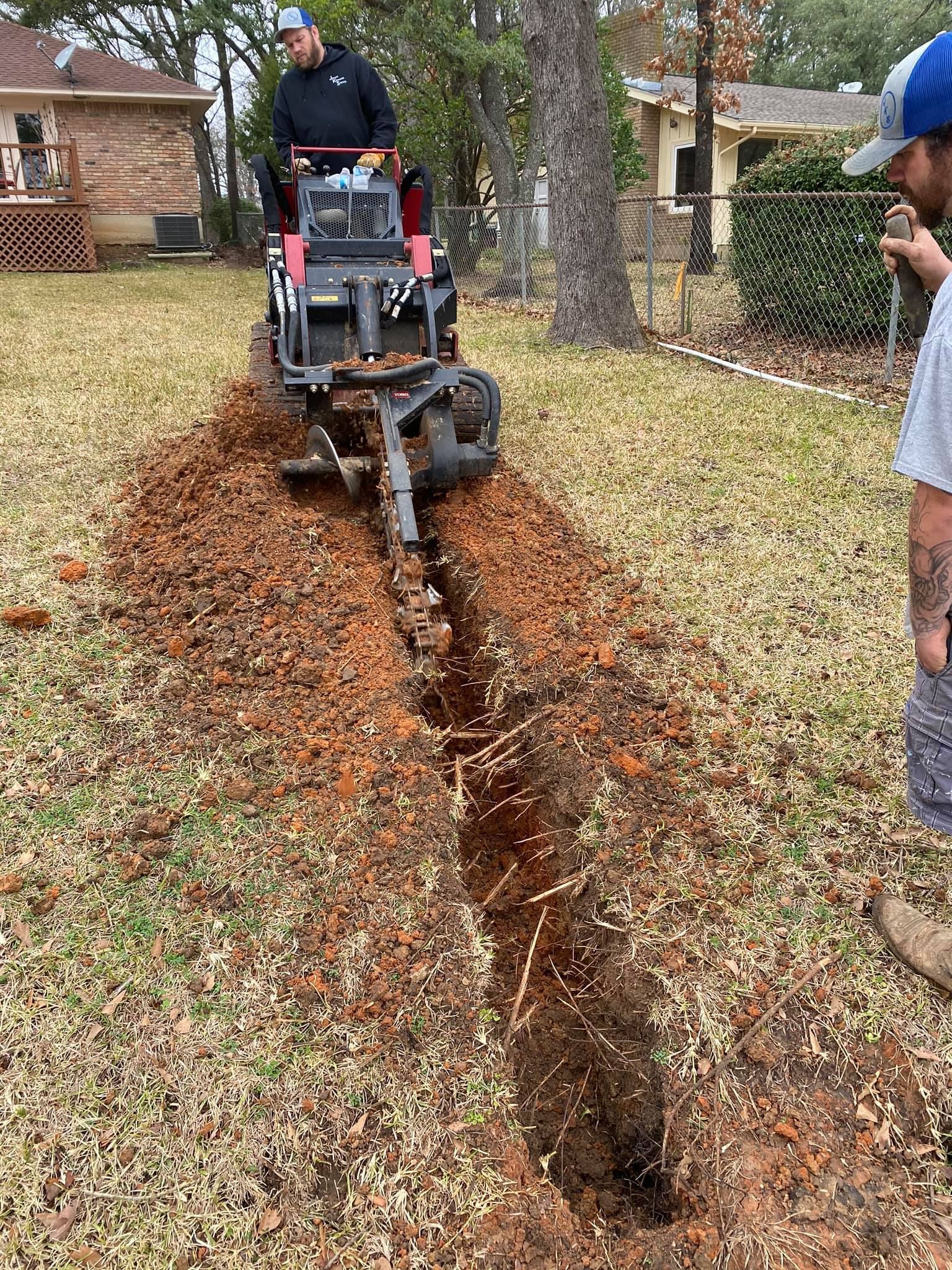 Man operating a trenching machine in a yard, creating a long trench in the ground. Another man watches.