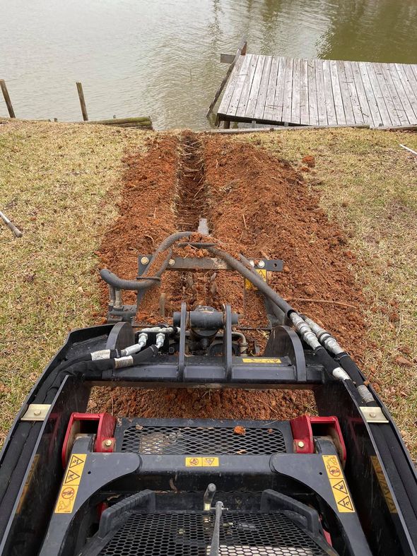 Bobcat machine spreading mulch along a grassy bank toward a lake and dock.