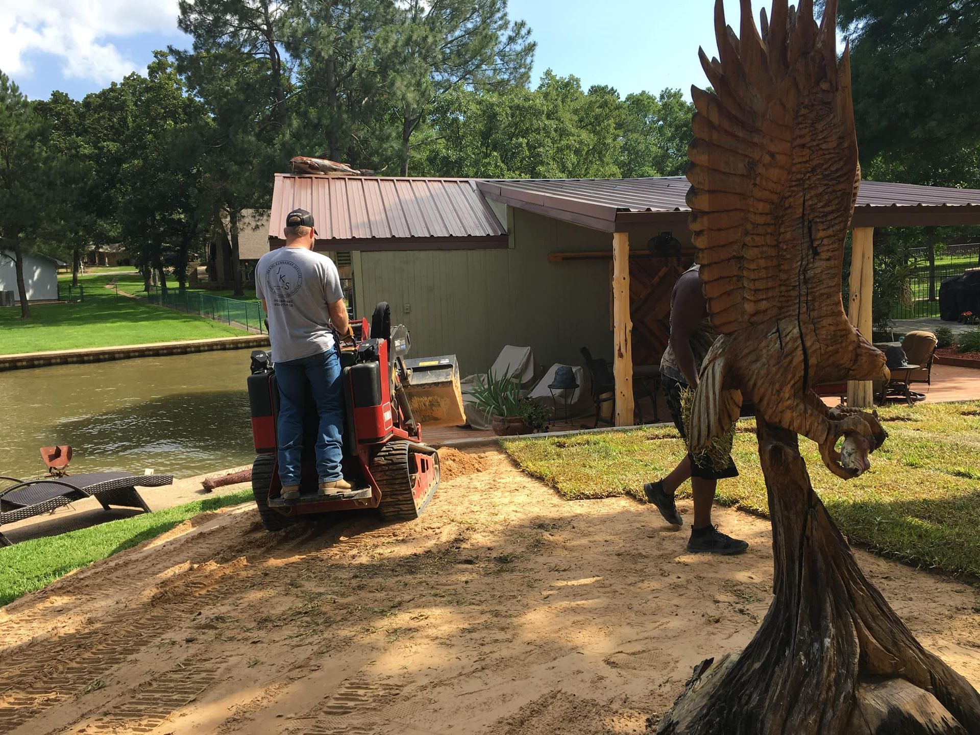 Man driving a small tractor, another man walking past large eagle sculpture. Landscaping project near pond, building.