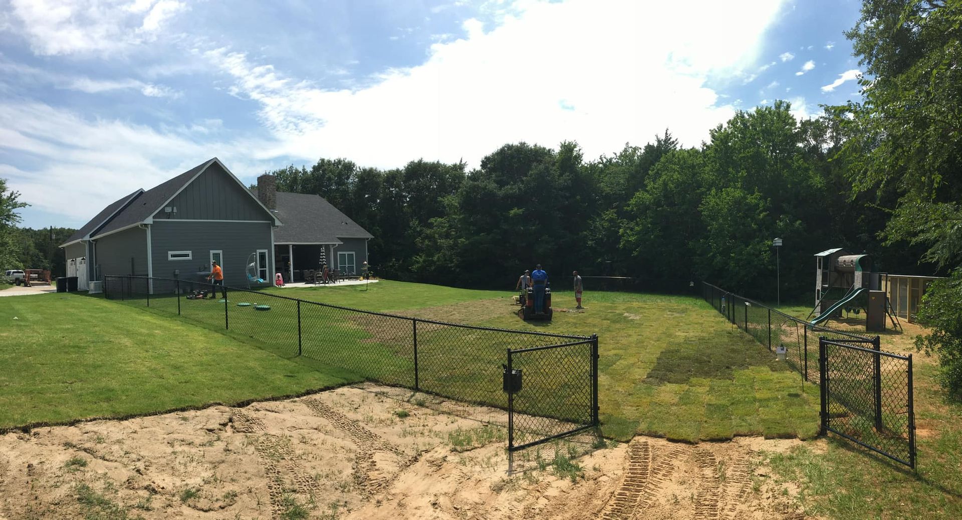 A black fenced backyard with a house, play area, and green grass under a blue sky.