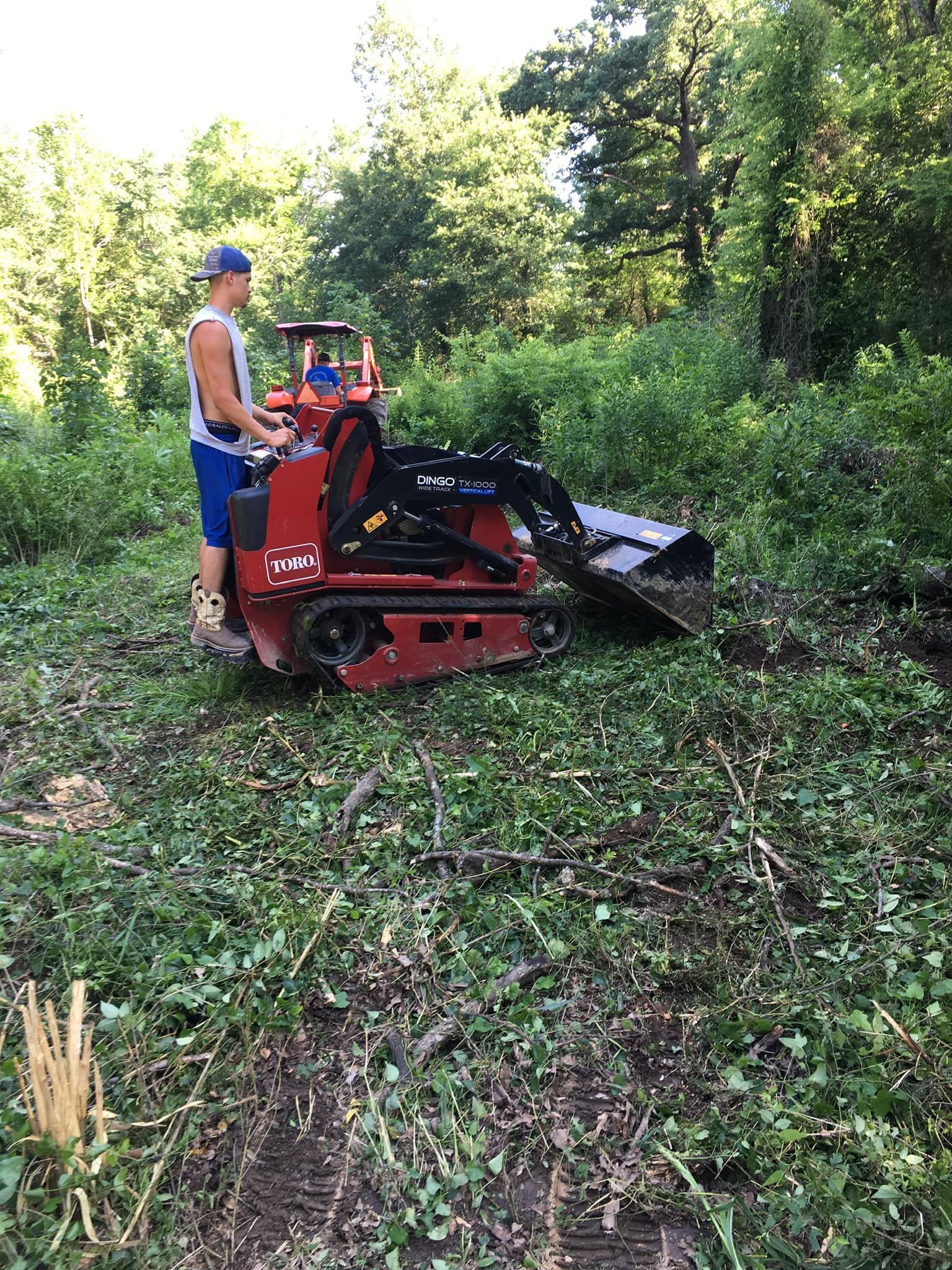 Man operating a red mini-skid steer with brush cutter attachment, clearing vegetation in a wooded area.