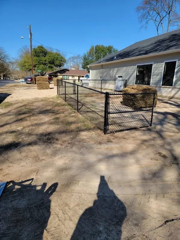 Black chain-link fence encloses a dirt area with hay bales near a building under a sunny sky.