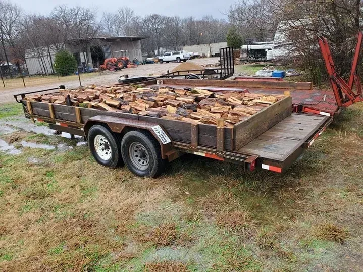 Trailer loaded with firewood on a grassy, overcast day. Buildings and trees in the background.