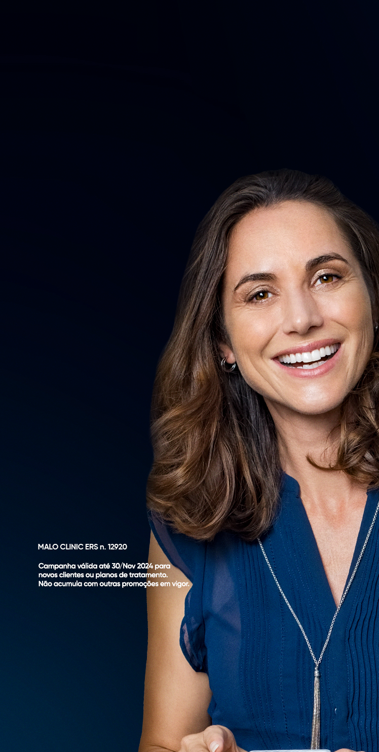 Retrato de uma mulher de meia-idade com um sorriso radiante, exibindo dentes brancos e bem cuidados. Ela tem cabelos castanhos ondulados até os ombros e veste uma blusa azul escura, com um fundo azul-marinho que realça sua expressão alegre e saudável.