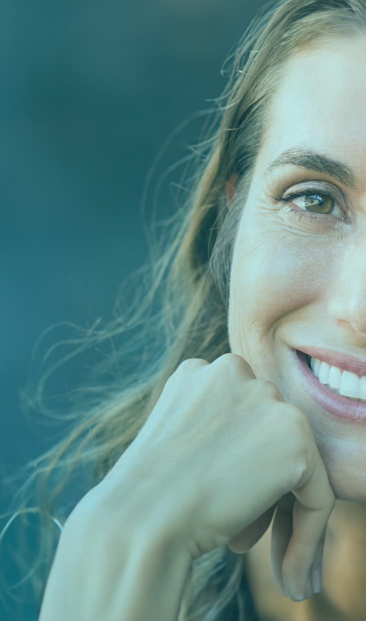 Retrato de uma mulher de meia-idade com um sorriso radiante, exibindo dentes brancos e bem cuidados. Ela tem cabelos castanhos ondulados até os ombros e veste uma blusa azul escura, com um fundo azul-marinho que realça sua expressão alegre e saudável.