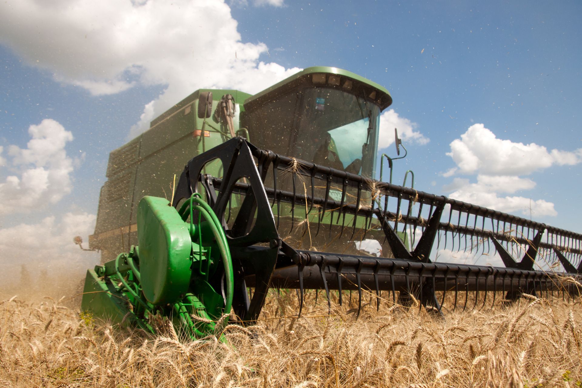 Green John Deere combine with black header. Outside a building, blue sky, harvest scene.