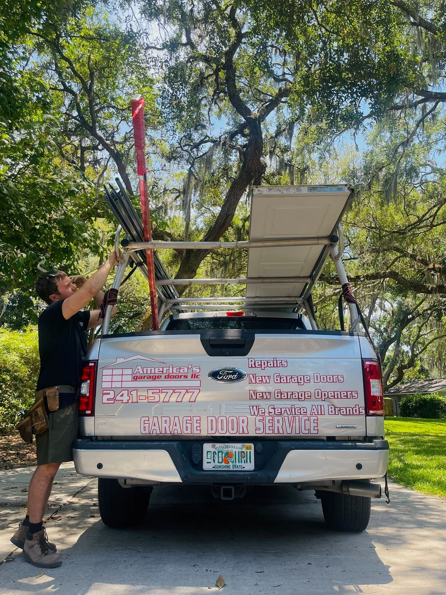 A technician in work attire secures a ladder and equipment onto the rack of a silver garage door service truck.