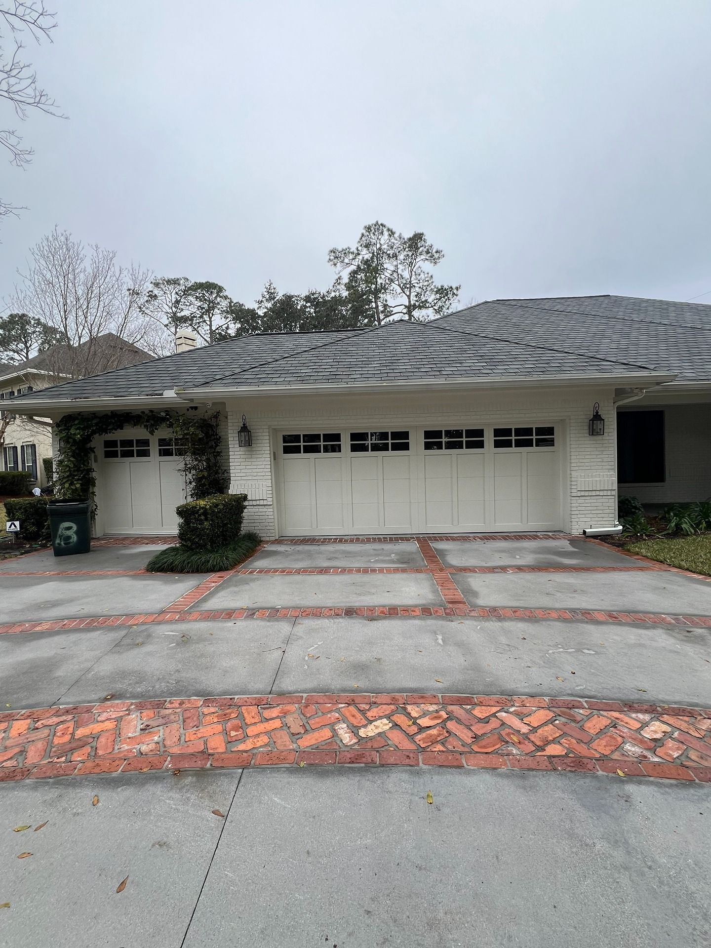 A light-colored house with three white garage doors and a brick-patterned concrete driveway under a cloudy sky.