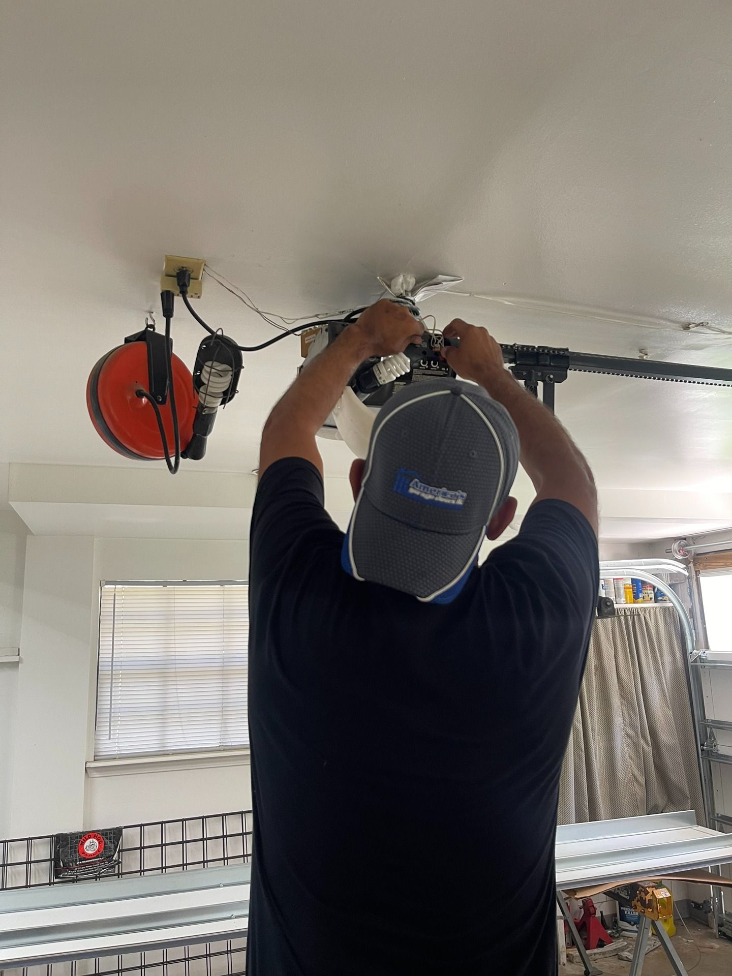 A technician in a gray cap works on a ceiling-mounted garage door opener in a garage.