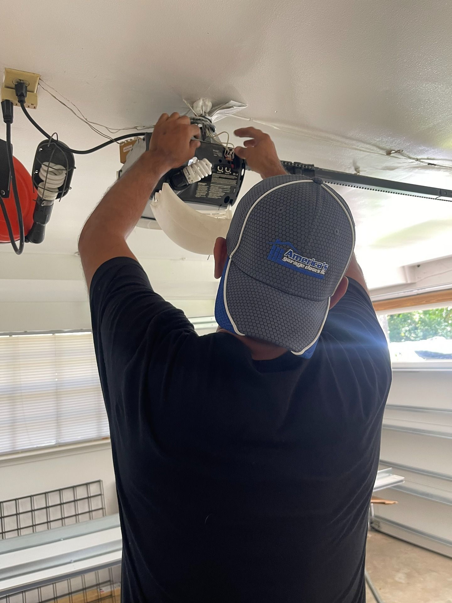 A technician in a branded cap works on a garage door opener mounted to a ceiling.