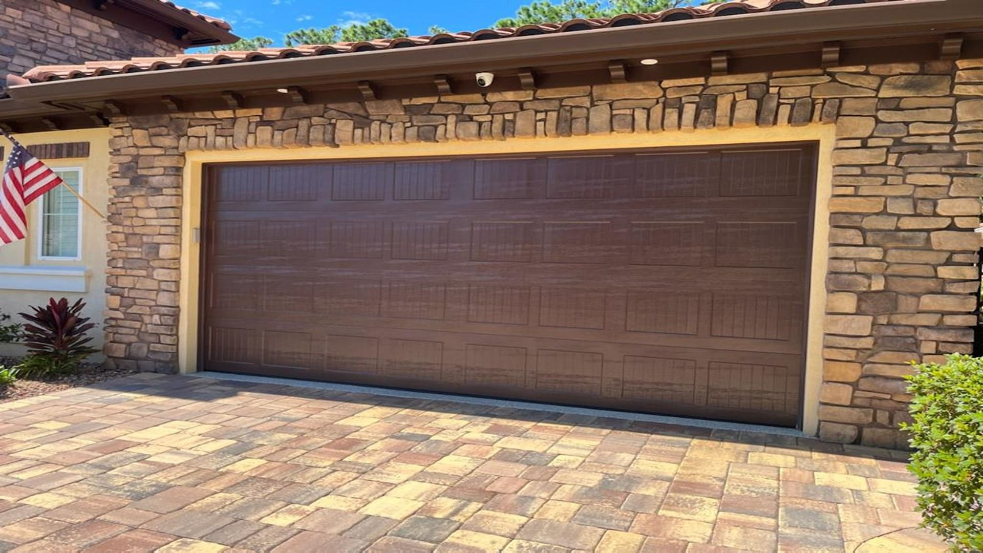 A brown garage door on a stone-facade house with a paved driveway and an American flag visible to the left.