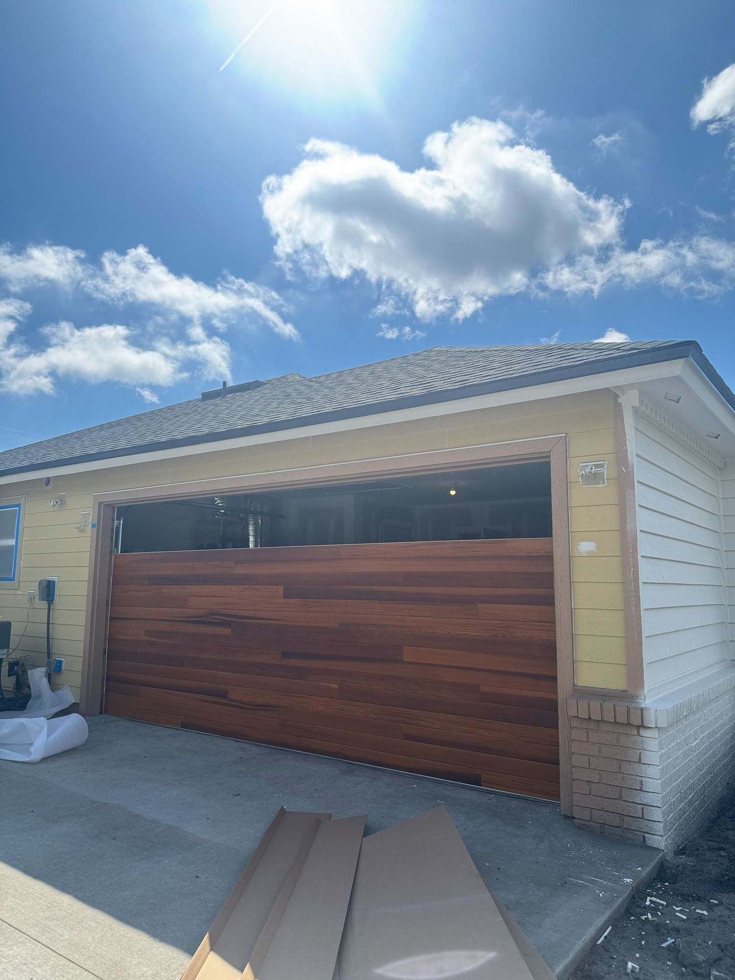 A partially installed horizontal wood-plank garage door on a yellow house under a sunny, blue, cloudy sky.
