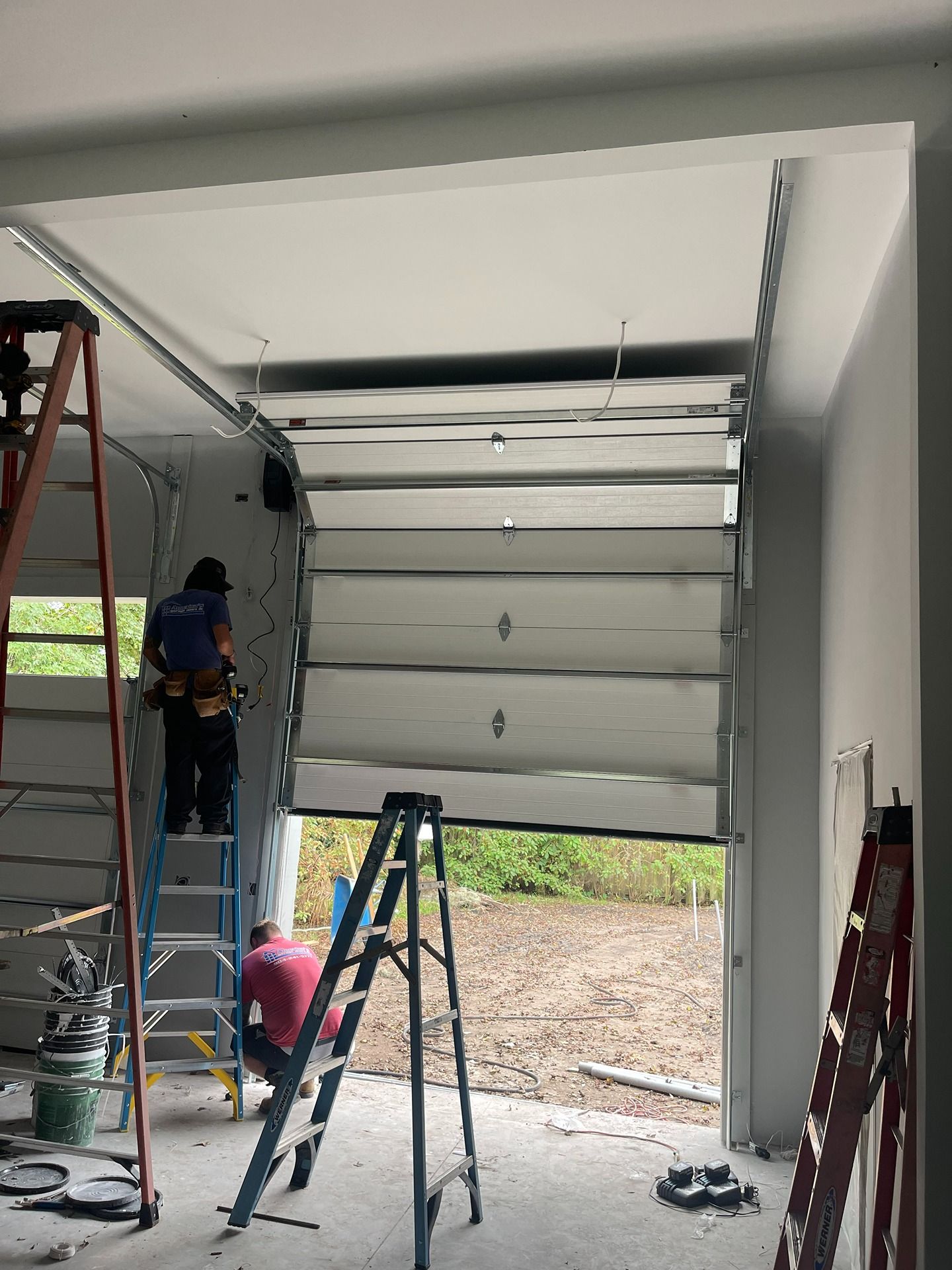 Two workers on ladders install a sectional garage door in an unfinished building.