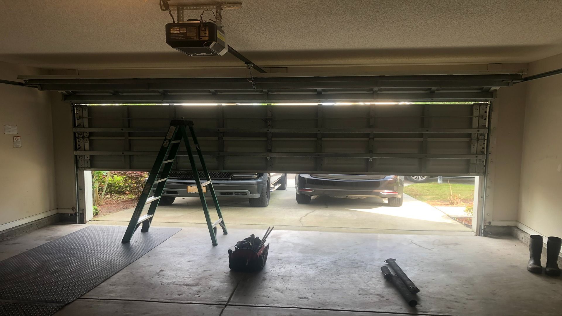 A partly opened garage door showing a driveway with parked cars, a step ladder, and tools on the garage floor.