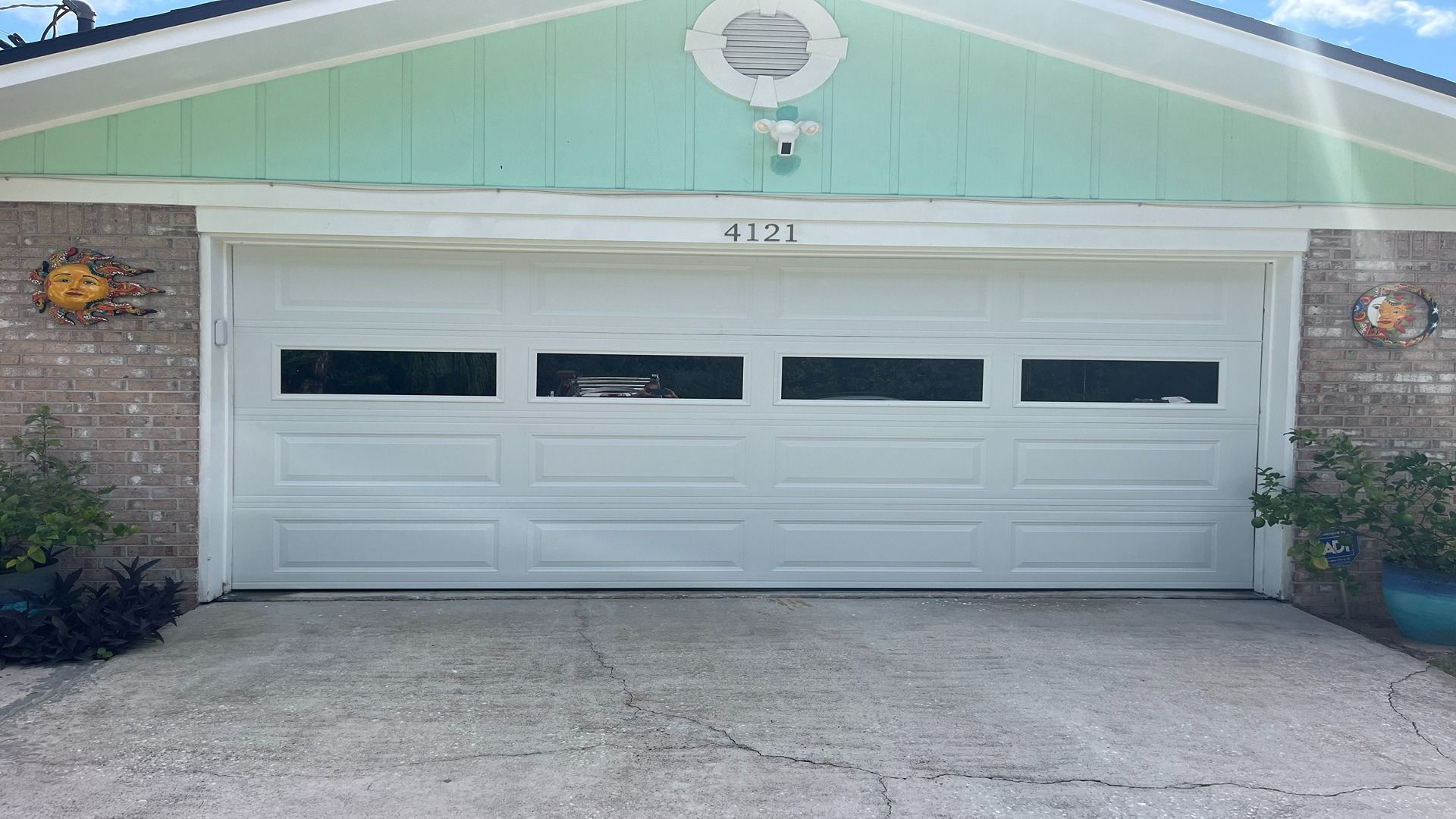 A white residential garage door with four rectangular windows, set in a brick house with light green siding above.