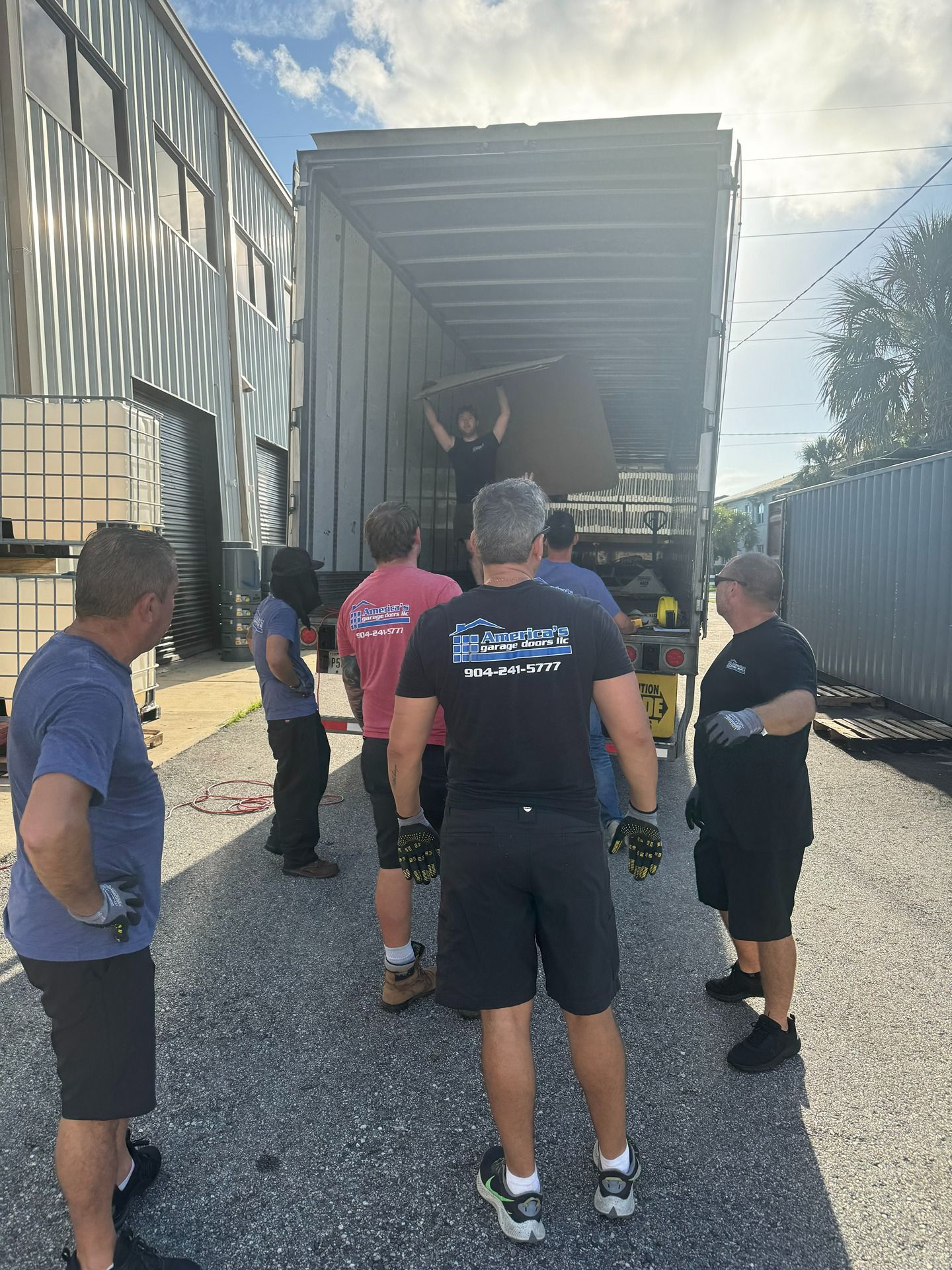 A group of people standing outside a semi-trailer truck, watching one person inside the trailer move a large object.