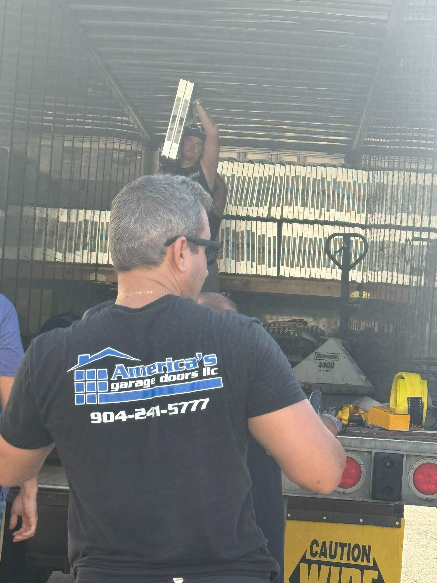 A person in a black company t-shirt stands before a trailer loaded with stacks of white garage door panels.