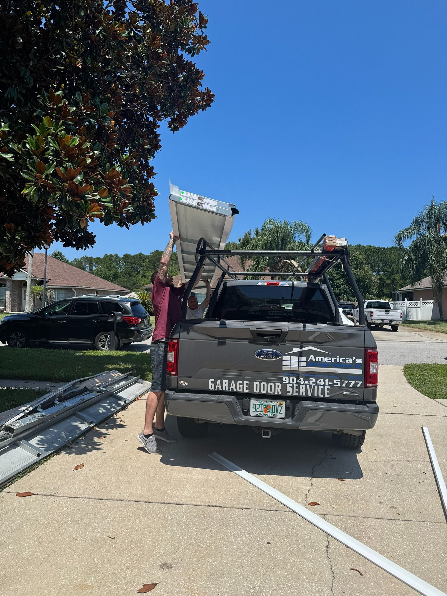 A technician loads a metal panel onto the rack of a grey pickup truck parked in a residential driveway.