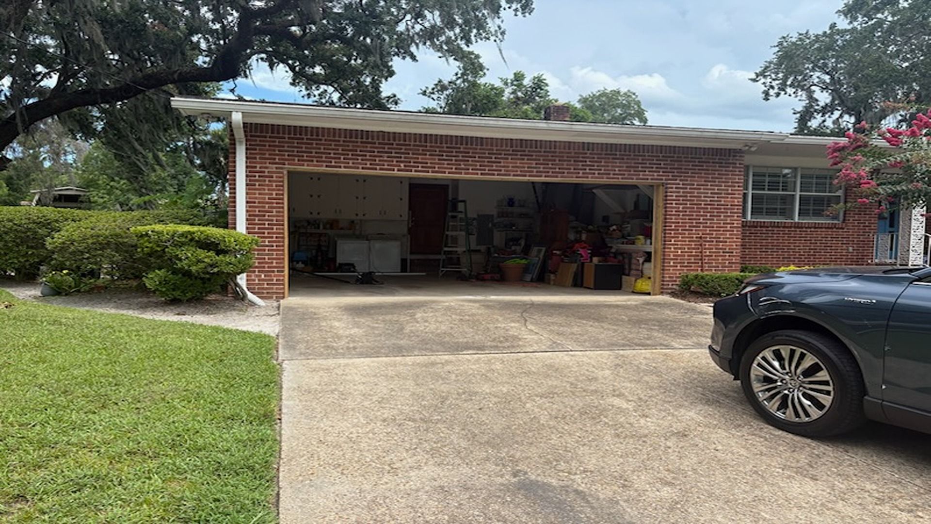 A brick house with an open garage, a concrete driveway, a green lawn, and part of a dark car on the right.