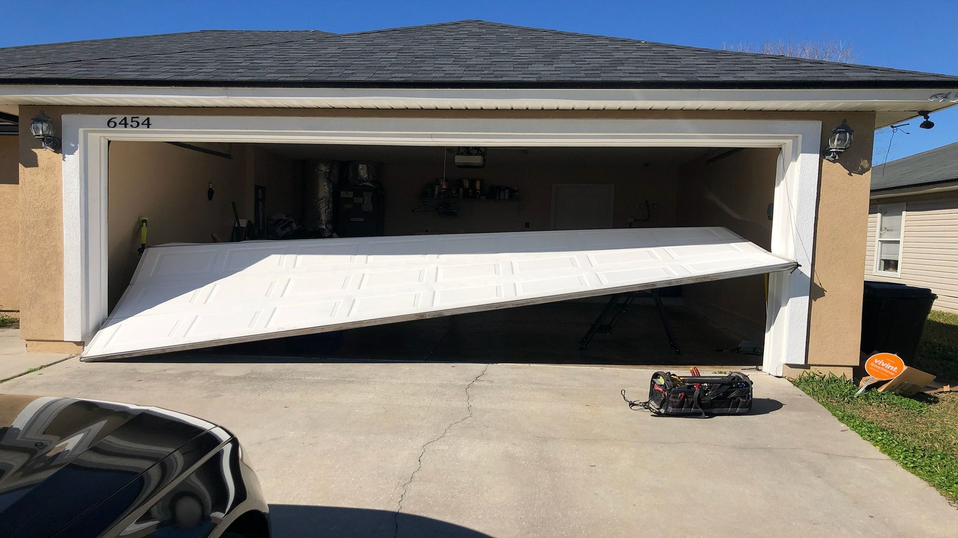 A white garage door hanging crookedly from its tracks on a beige residential house.